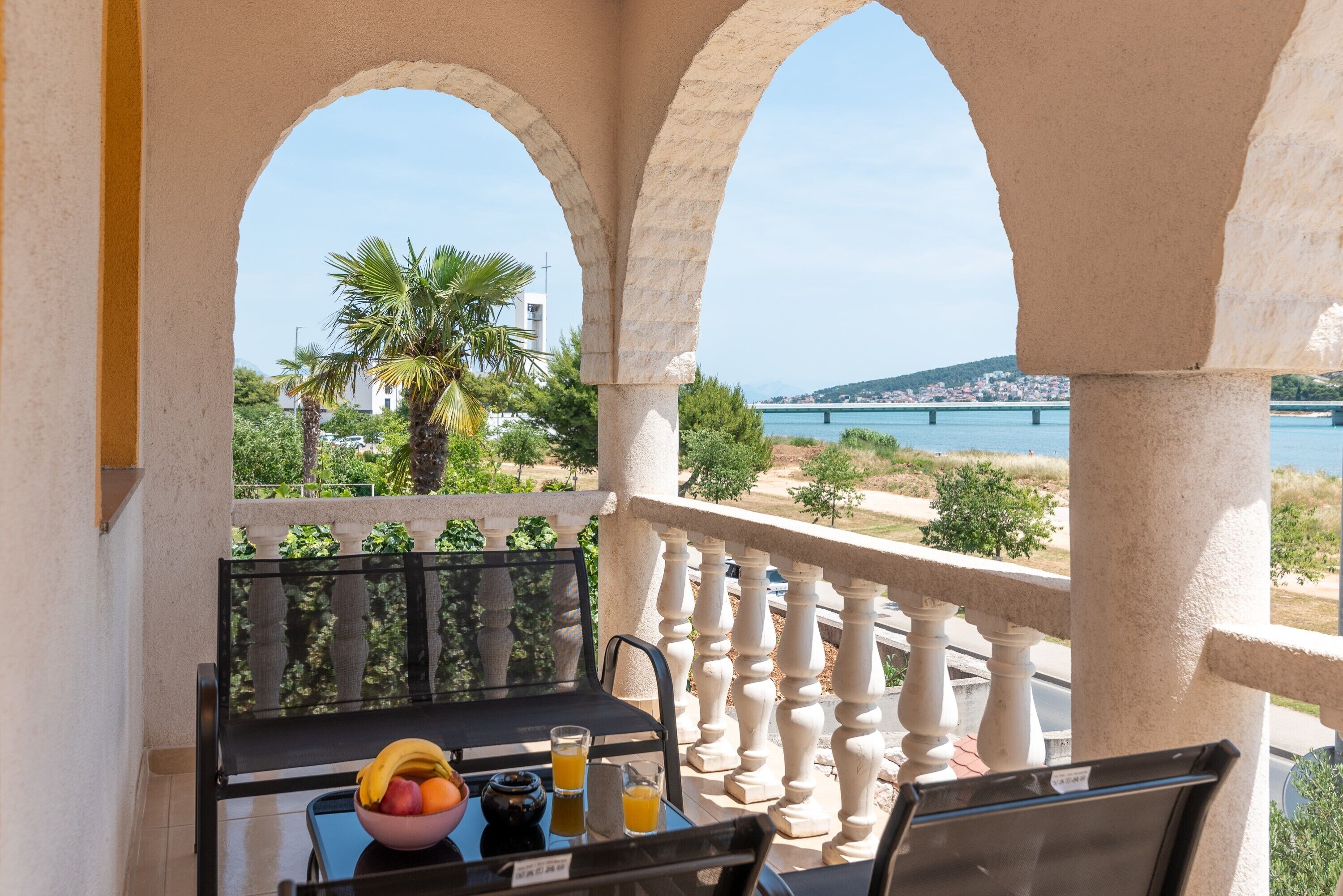 A balcony with a table set for breakfast, overlooking a coastal view with palm trees and a bridge.