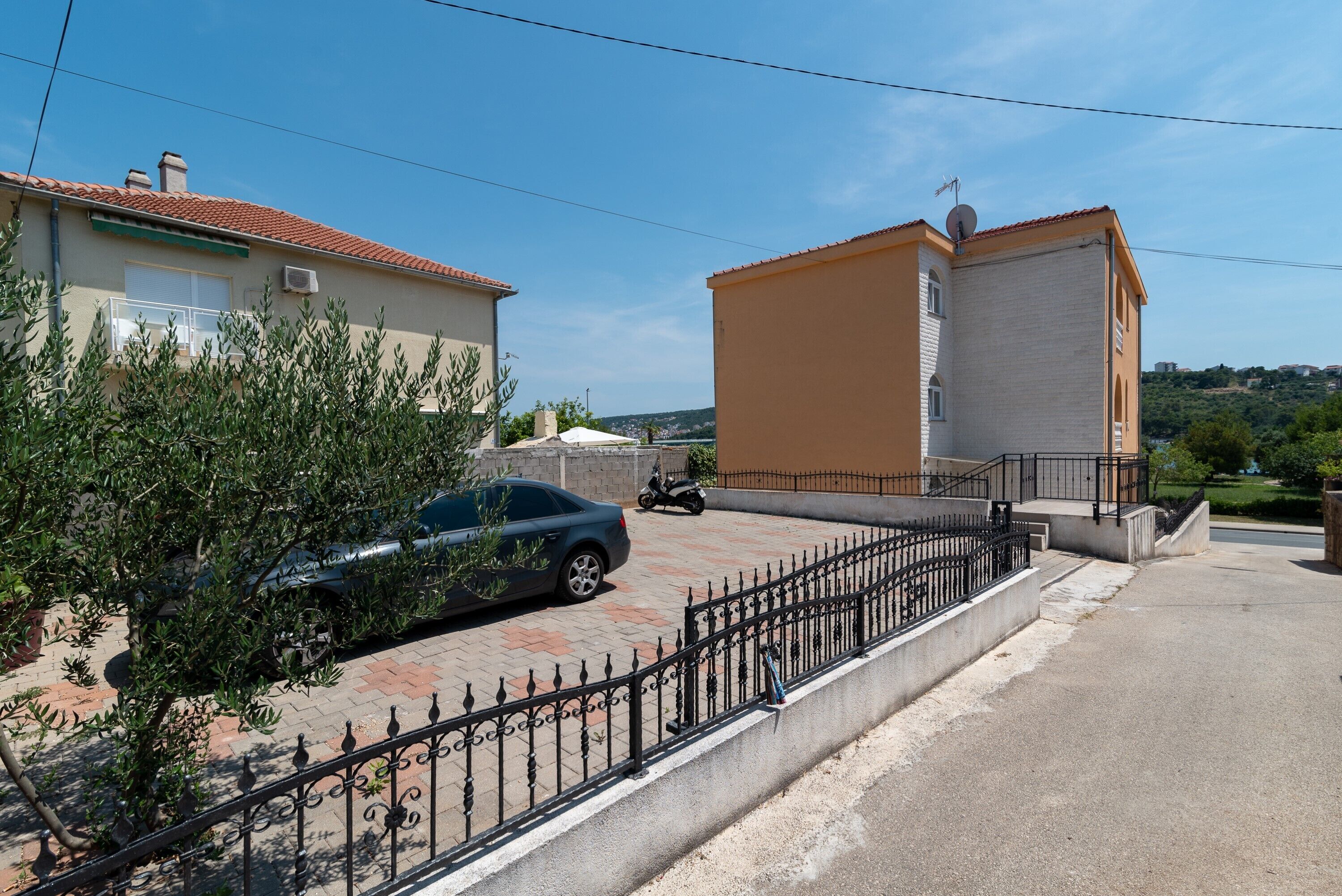 Il s’agit d’un quartier résidentiel avec une allée pavée, une voiture garée et une moto. On y trouve deux bâtiments aux styles architecturaux distincts : l’un est doté d’un toit en tuiles, l’autre présente une façade plate.