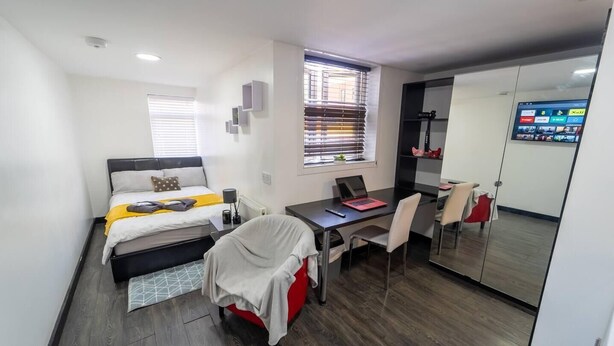 Habitación de hotel compacta con cama, escritorio con laptop, silla, mesita pequeña y espejo.