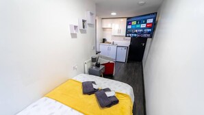 Habitación de hotel pequeña y funcional con cama, un escritorio, kitchenette y televisión que muestra varios canales.