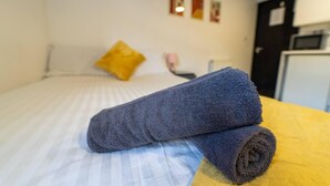 Ein ordentlich bezogenes Bett mit einem aufgerollten Handtuch und einer gelben Decke, einem gelben Kopfkissen und einem kleinen Nachttisch mit einer pinkfarbenen Lampe.