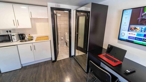 A modern kitchen with white cabinets, a microwave, and a coffee maker. There is a laptop on a desk with a TV displaying Netflix options. - Modern studio available close to the city (Birmingham)
