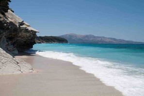 A beach with clear turquoise waters, a rocky cliff, and a distant mountain range.