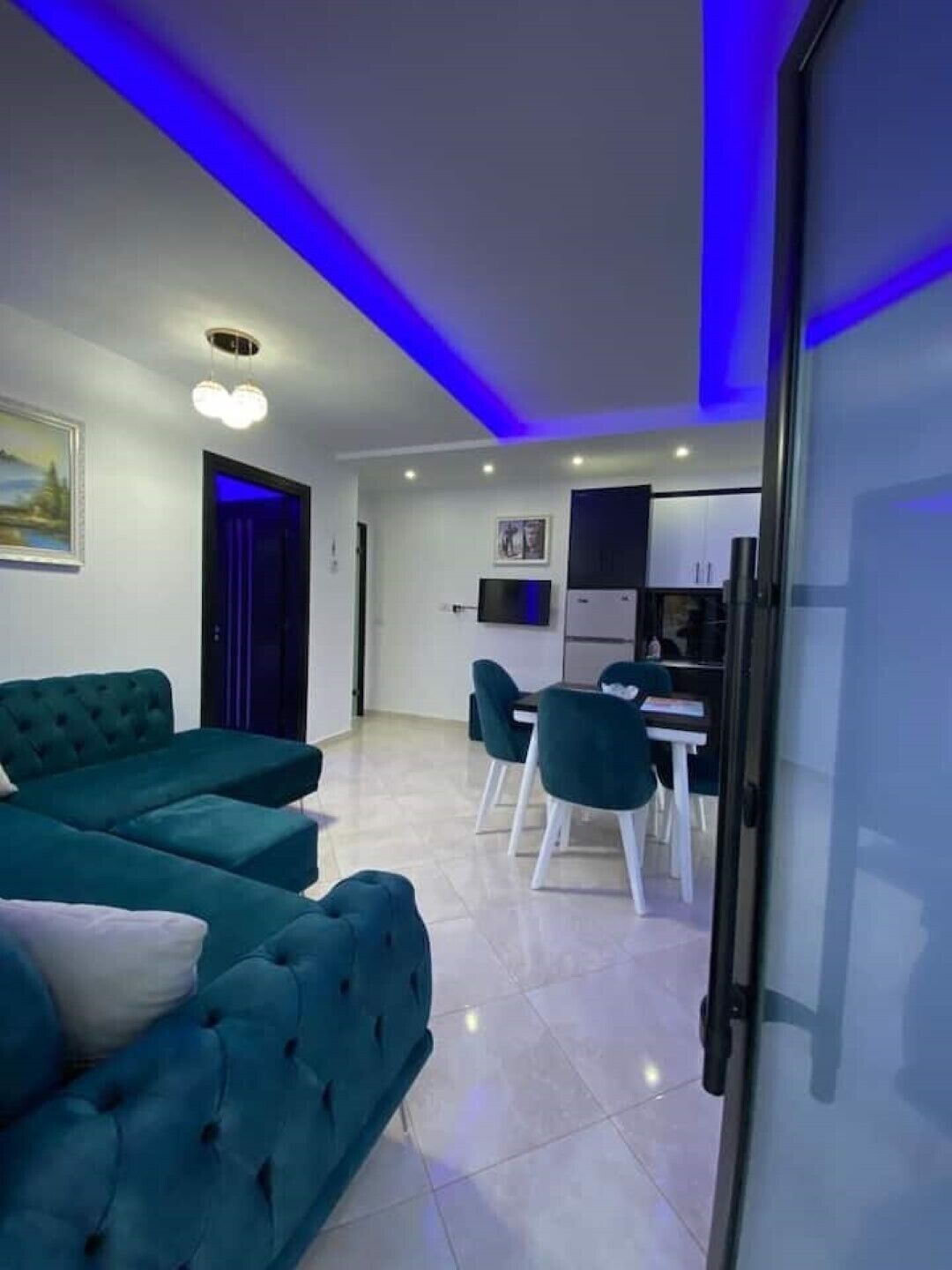 A modern living room with a teal sofa, a dining area with a table and chairs, and a kitchen in the background.