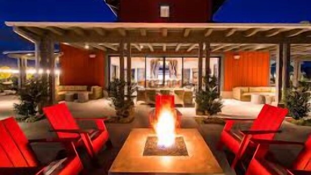 Een overdekt terras met een centraal vuurkorf, rode stoelen en een rood gebouw op de achtergrond.