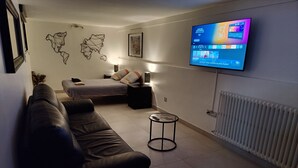 A modern living room with a leather sofa, a small round table, a bed with pillows, a TV mounted on the wall, and a radiator. - L'escapade Enchantée Paris-disneyland (Noisy-le-Grand)