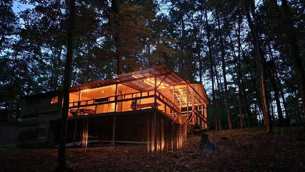 A modern wooden cabin with a glass facade, surrounded by a forest.