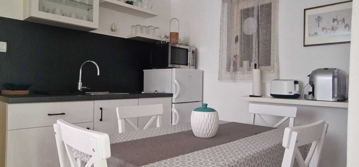 A compact kitchen with a microwave, refrigerator, and toaster on the counter. There are white chairs around a table with a patterned tablecloth.