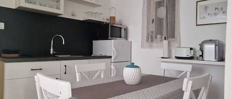A compact kitchen with a microwave, refrigerator, and toaster on the counter. There are white chairs around a table with a patterned tablecloth.