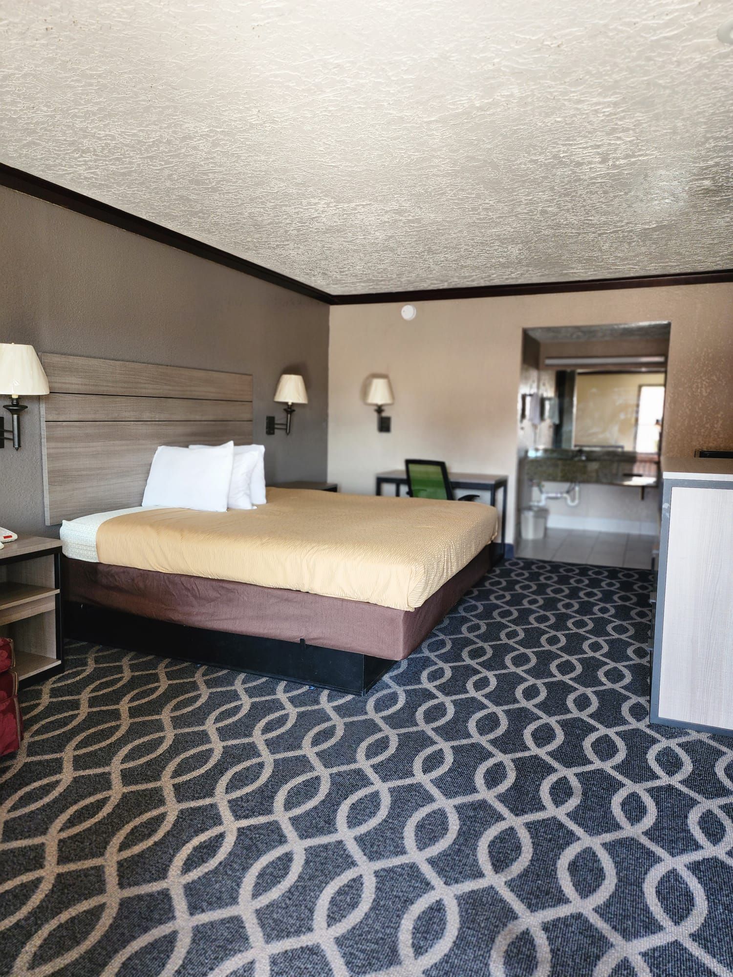 A hotel room with a bed, bedside table, lamp, and a kitchenette area.