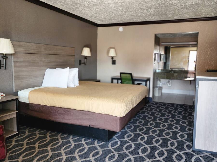 Habitación de hotel con cama, mesita de noche, lámpara y una pequeña cocina.