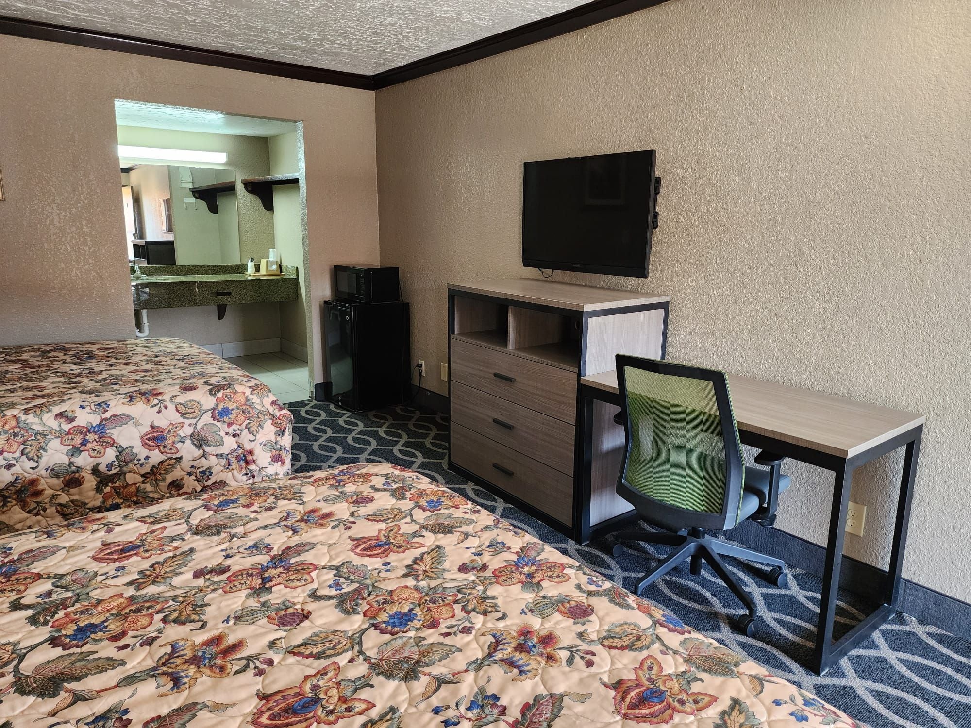 Quarto de hotel com cama, escrivaninha, cadeira, televisão e pequeno espaço de cozinha.