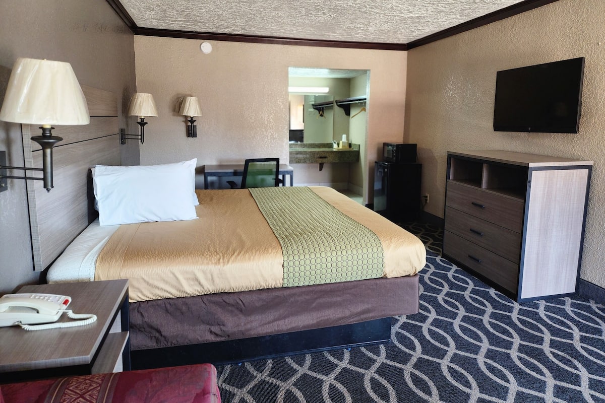 A hotel room with a bed, bedside table, lamp, TV, and a desk with a telephone.