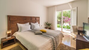 A bedroom with a wooden headboard, a bed with a patterned bedspread, a bedside table with a lamp, a TV stand, and a view of a garden through a sliding glass door. - Gumonca in Supetar With 6 Bedrooms and 5 Bathrooms (Supetar)