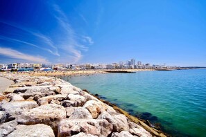 Un paisaje urbano costero con un litoral rocoso, una playa de arena y una hilera de edificios al fondo.