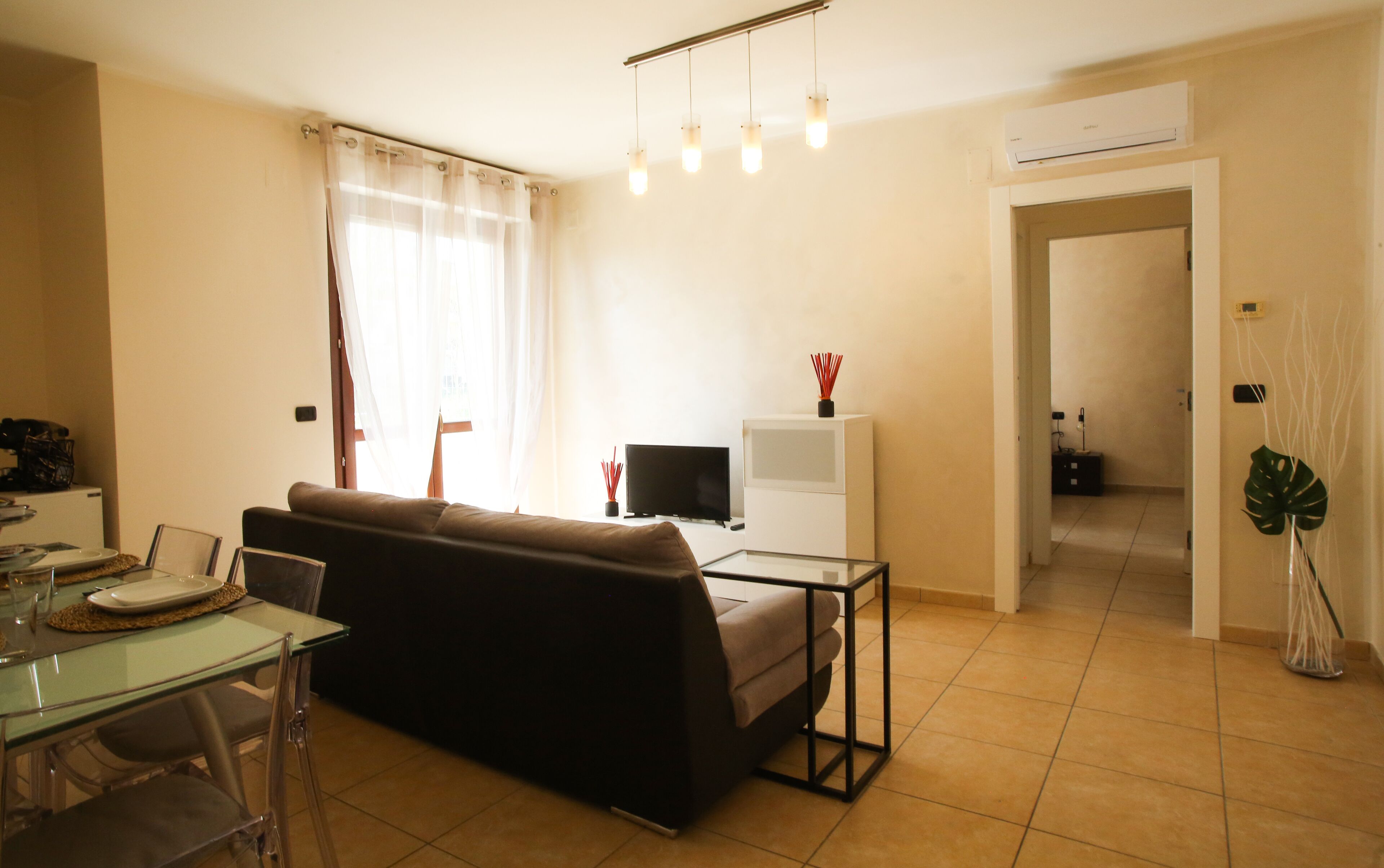 Una sala de estar con un sofá, una mesita de centro de vidrio, un televisor y un mueble blanco pequeño.