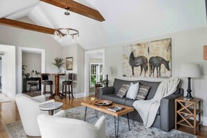 A spacious living room with a high ceiling, a wooden ceiling beam, a grey sofa, a coffee table, and a painting of horses.