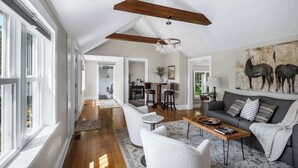 A spacious living room with a high ceiling, wooden beams, a chandelier, a grey sofa, and a large painting of cows. - Pet Friendly-Fenced Yard-Near Downtown (Skaneateles)