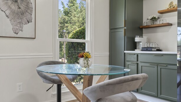A modern kitchen with a glass table, grey upholstered chairs, and a view of trees through a large window. - Spacious & Cozy ~ Close to Fort Moore ~ Pool Table (Columbus)