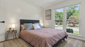 A bedroom with a bed, bedside lamps, a small table with a plant, and a large window with a view of trees and a car. - Spacious & Cozy ~ Close to Fort Moore ~ Pool Table (Columbus)