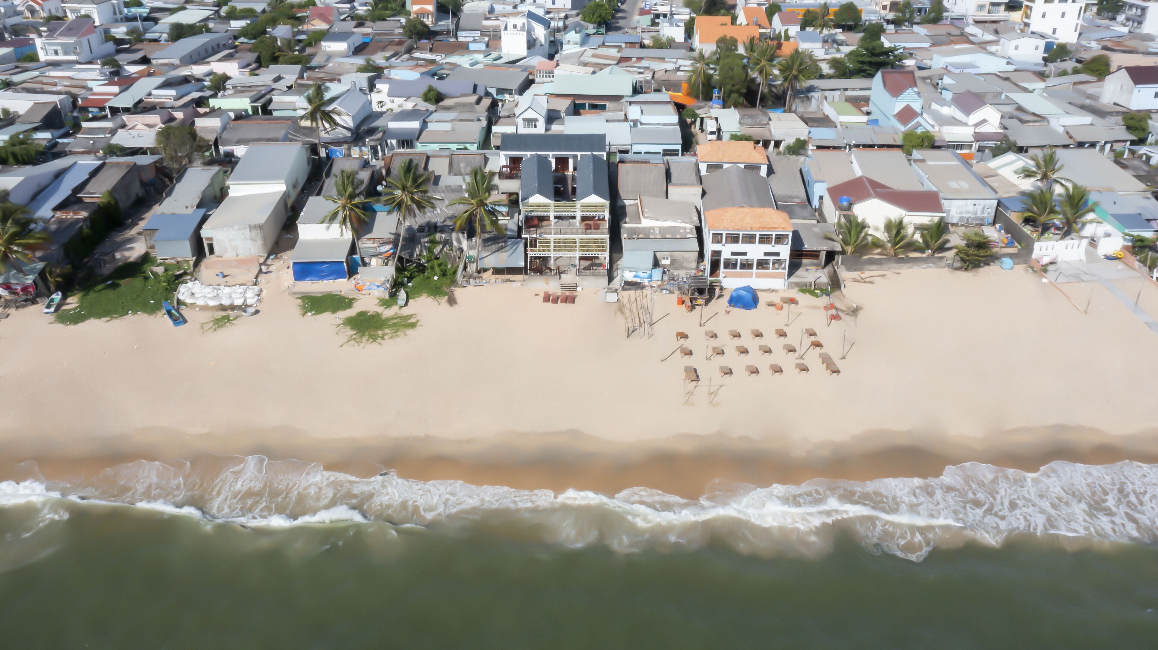 An aerial view of a coastal area with a sandy beach, a row of houses, and a building with a balcony overlooking the beach.