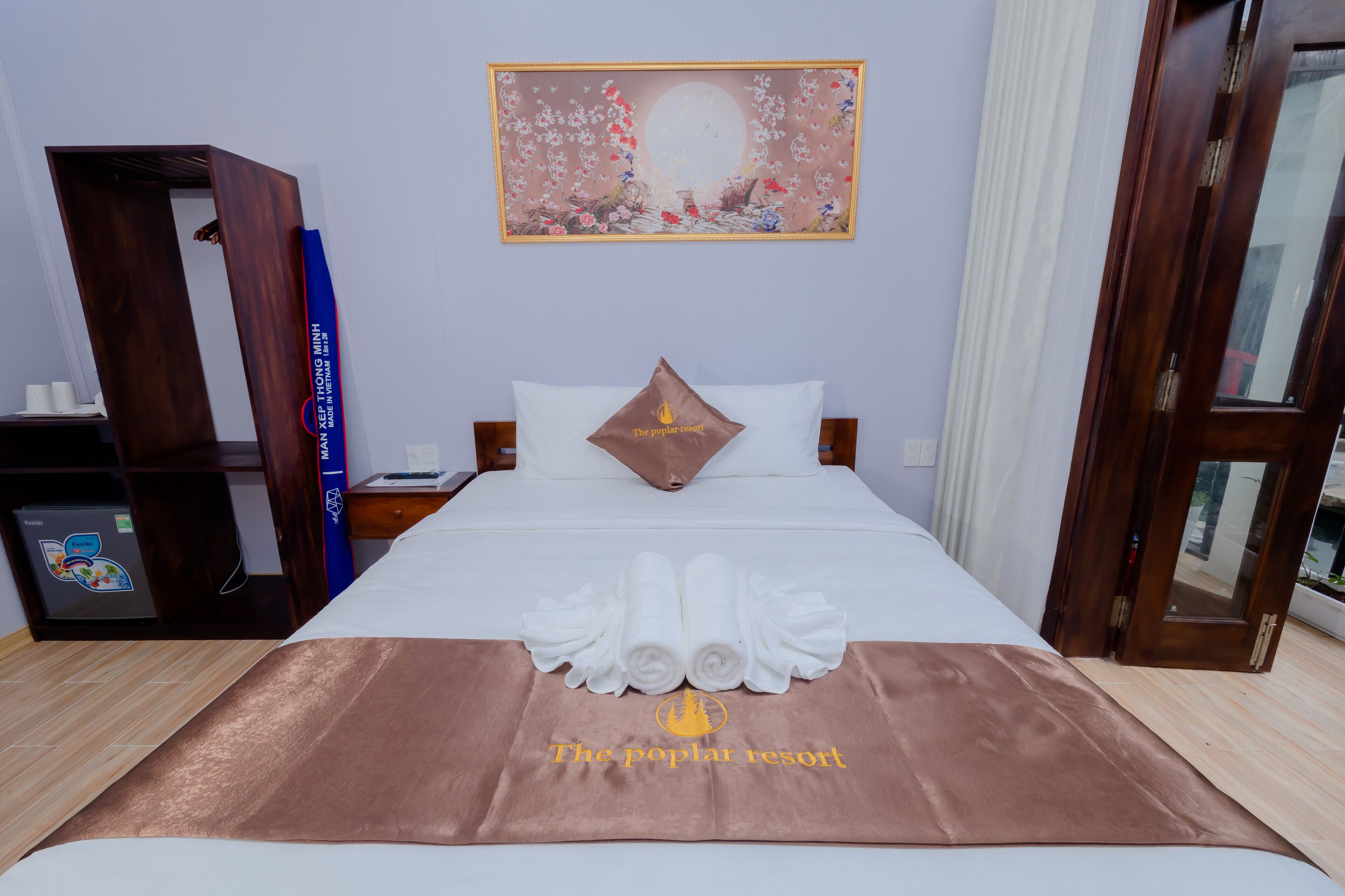 Une chambre d’hôtel avec un lit parfaitement fait, une couverture marron ornée du logo du complexe hôtelier, un oreiller à motif de flamme, une table de chevet, une armoire en bois et un tableau accroché au mur.