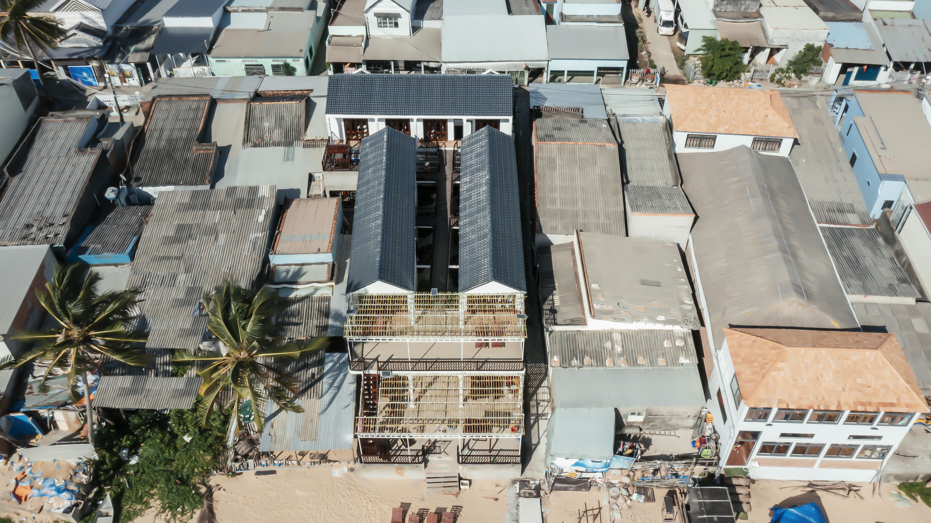 An aerial view of a residential area with multiple houses, some with corrugated metal roofs, and a few with tiled roofs. There are palm trees and a sandy ground visible.