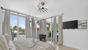 A bedroom with a large bed, a TV mounted on the wall, and a view of the outdoors. - Coastal Condo Retreat: 3BR 2.5BA w/ Heated Infinity Pool (Deerfield Beach)