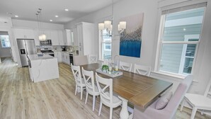 A modern kitchen with white cabinetry, a central island, and a dining area with white chairs and a wooden table. - Private Heated Pool | FREE Golf Cart | 5 BR Home | 5 Outdoor Spaces | Near Dune Allen Beach (Santa Rosa Beach)