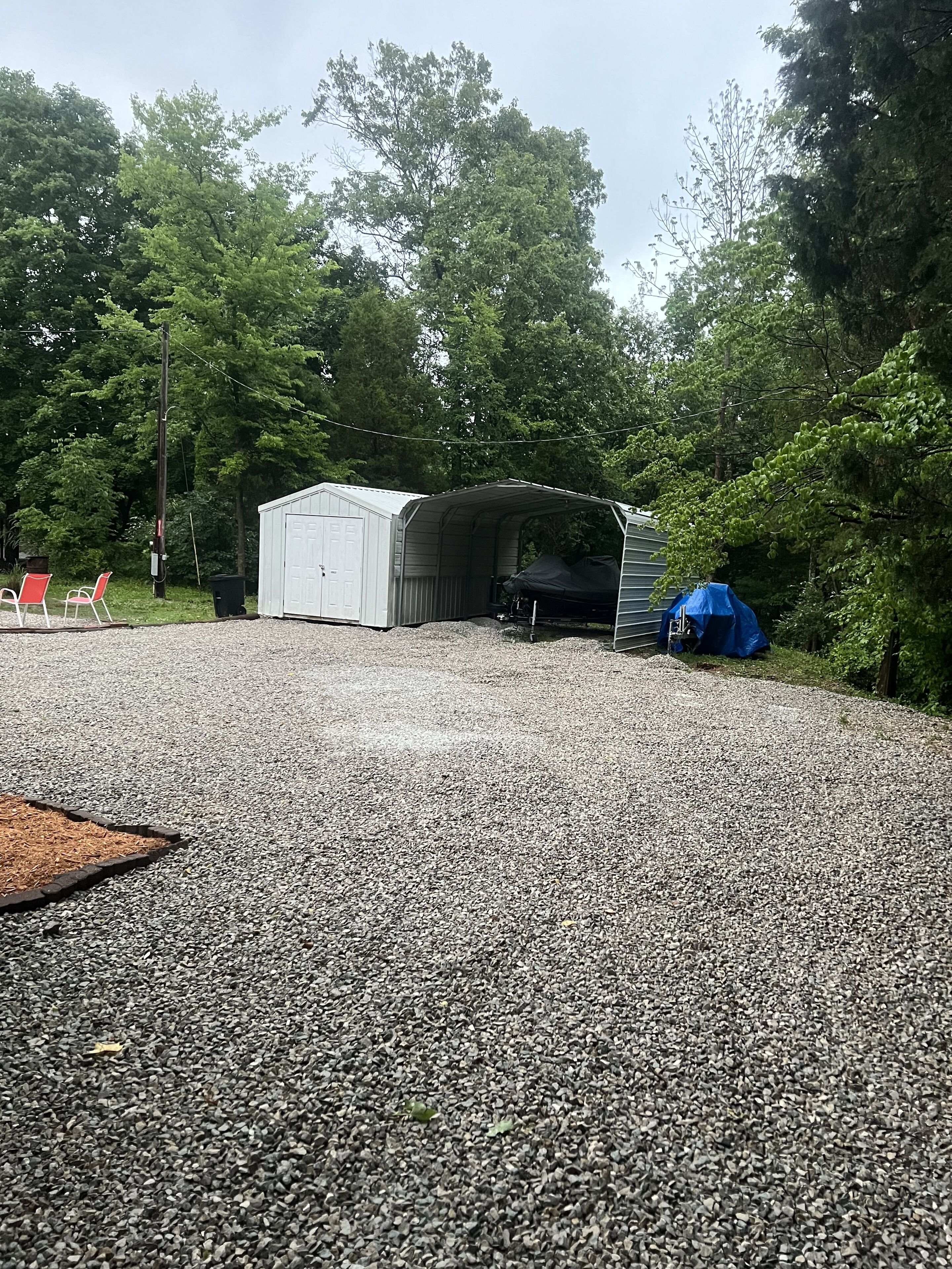 Ein schotterbedeckter Bereich mit einer überdachten Konstruktion, möglicherweise ein Schuppen oder eine Garage, und einer blauen Plane auf dem Boden.