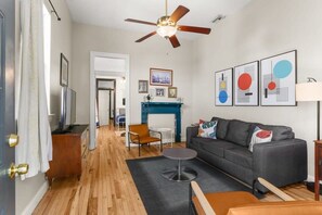 A modern living room with a grey sofa, wooden flooring, a ceiling fan, and framed artwork on the wall.