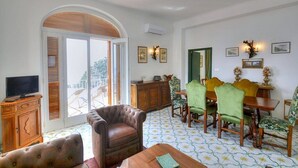 Un salon spacieux doté d’une grande fenêtre en arc de cercle, d’un téléviseur, d’un fauteuil en cuir, d’un coin repas avec une chaise recouverte d’un tissu vert et d’un sol à motifs.