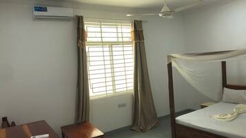Une chambre à coucher comprenant un lit, un bureau, une chaise, un ventilateur de plafond et une fenêtre avec des rideaux.