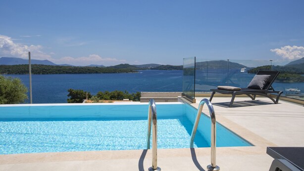 Piscina azul cristalina con barandilla de vidrio, una silla de descanso y vistas al mar y las colinas.