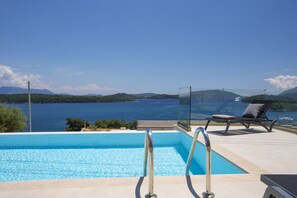 A clear blue pool with a glass railing, a lounge chair, and a view of the sea and hills. - Villa Orama Onorama in (Lefkada)