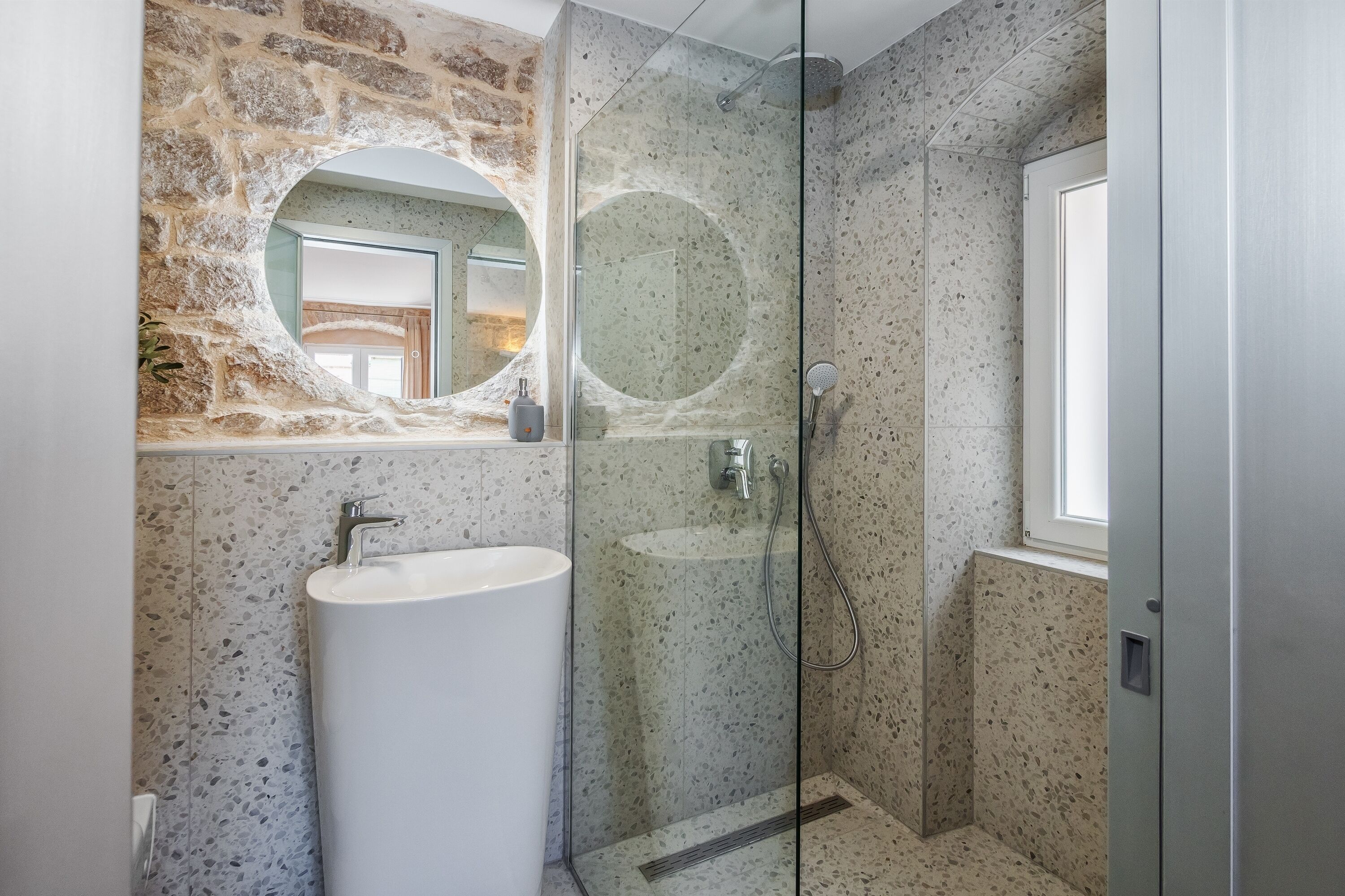 A bathroom with a stone wall, a round mirror, a sink, and a glass-enclosed shower.