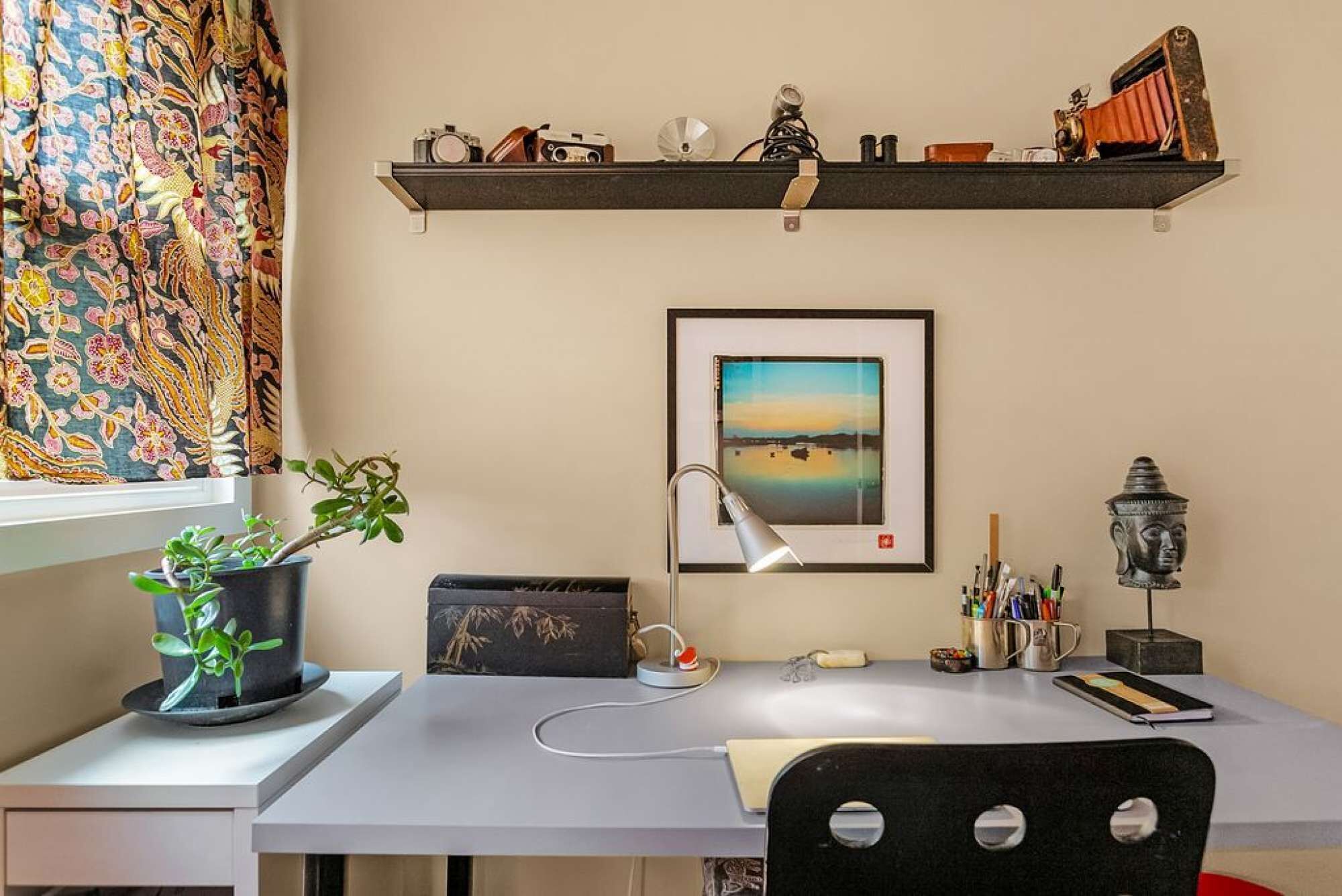 Ein Arbeitsplatz mit Schreibtisch, Stuhl, Topfpflanze, Lampe und einem gerahmten Bild an der Wand.