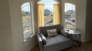 A living room with a gray sofa, a small round table, and three large windows with curtains.