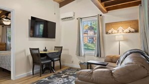 A cozy living room with a sofa, a dining area with chairs, a TV mounted on the wall, and a window with curtains. - Cozy Woodland 1BR Cottage Near Trails (Hendersonville)
