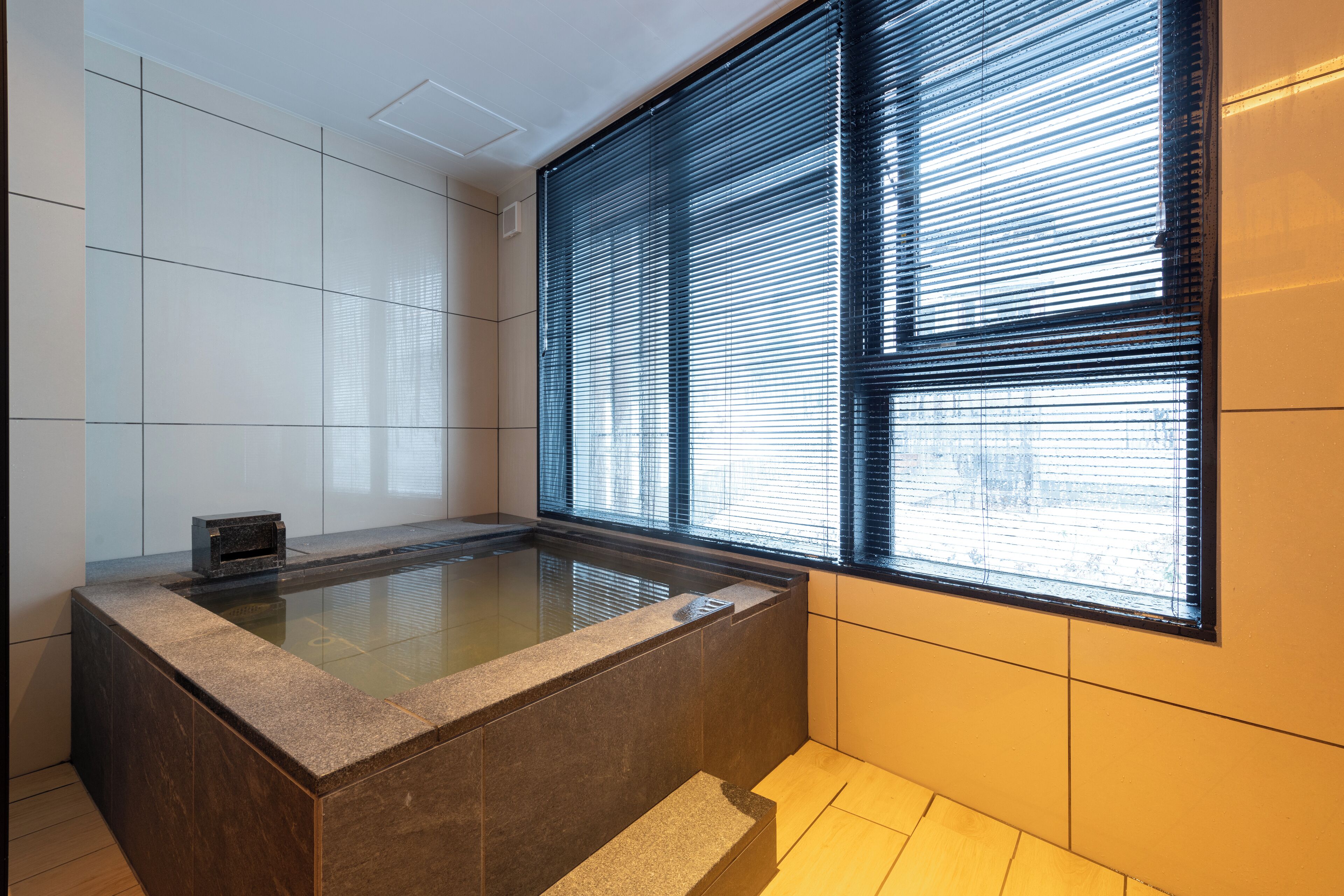 A modern bathroom with a freestanding bathtub, large windows with blinds, and tiled walls.