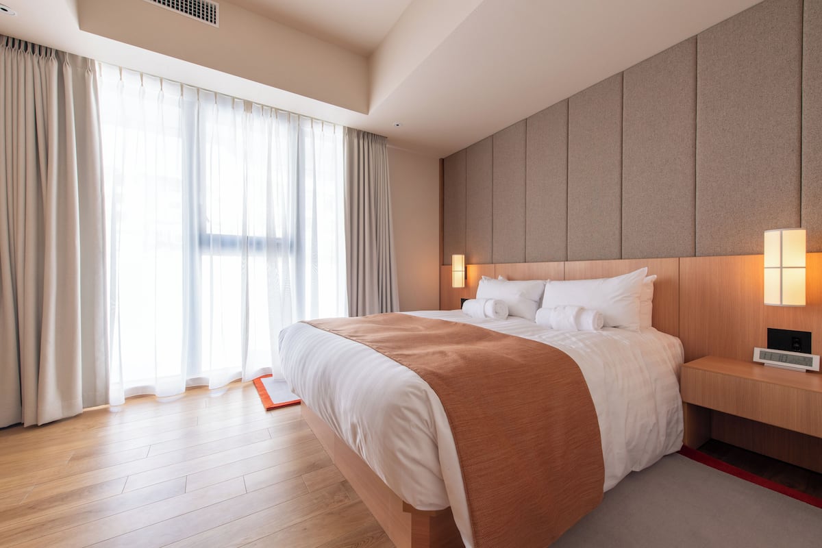 A modern hotel room with a large bed, wooden flooring, a headboard with built-in lighting, and a view of the outdoors through sheer curtains.