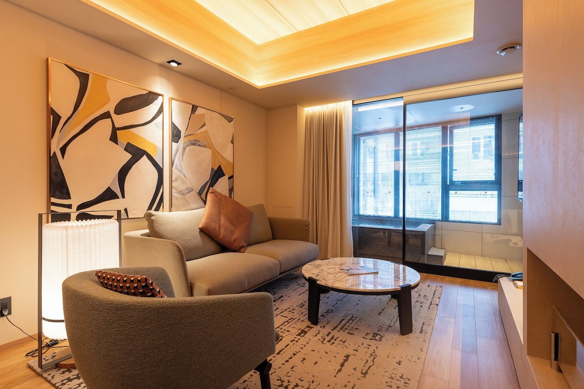 A modern living room with a sofa, armchair, coffee table, abstract wall art, and a large window with blinds.