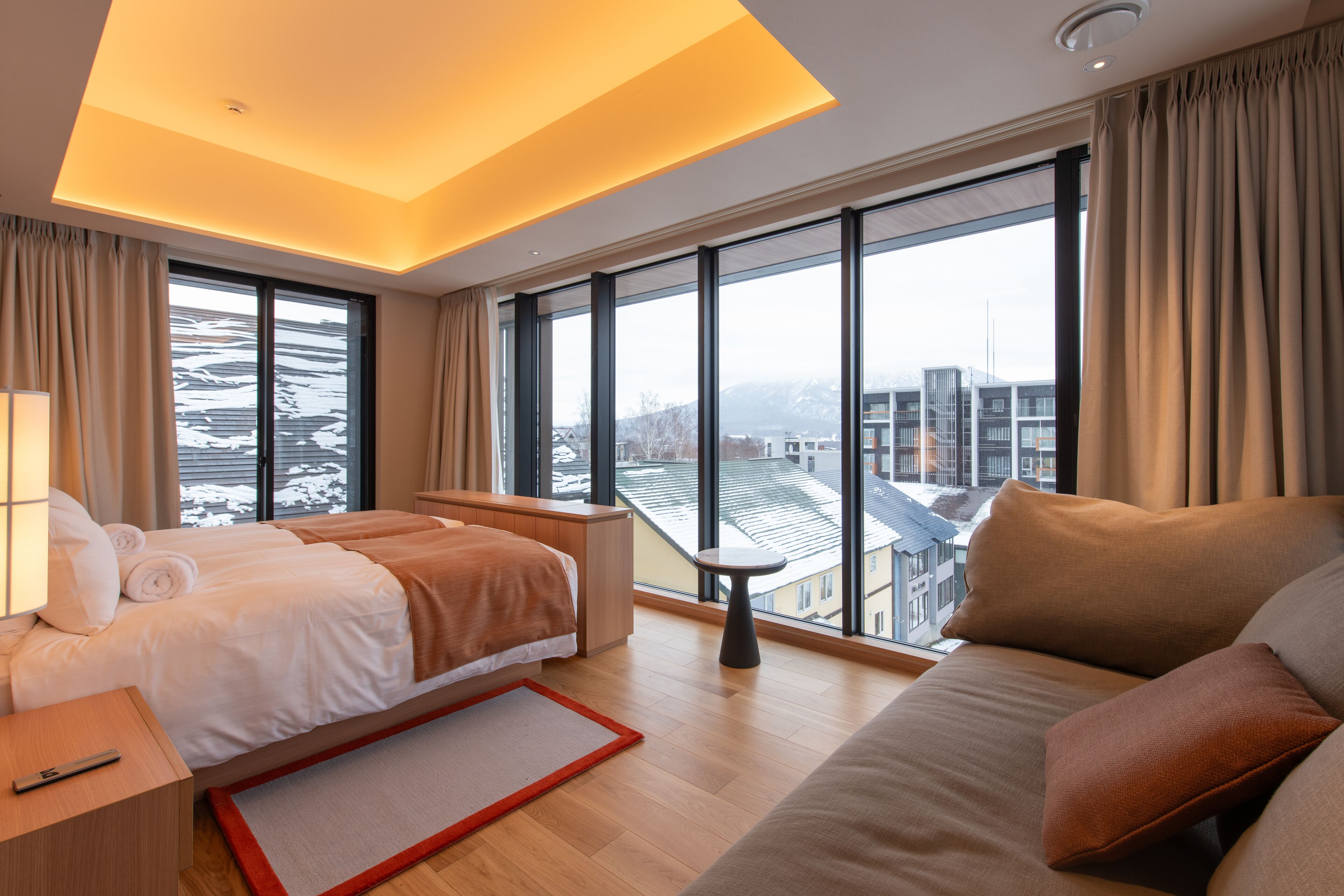 A modern hotel room with a large bed, a sofa, and a view of the cityscape through large windows.