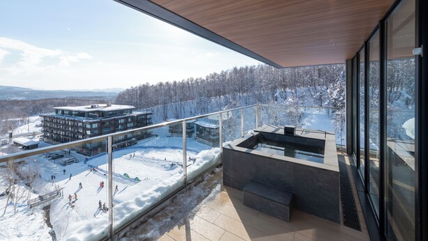 Un bâtiment moderne doté d’un balcon en verre donnant sur un paysage enneigé et une piste de ski.