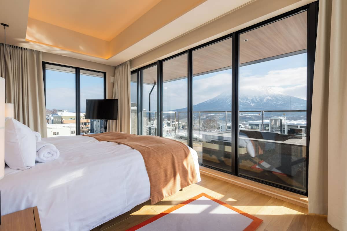 A hotel room with a large bed, a flat-screen TV, and a view of a mountain through floor-to-ceiling windows.