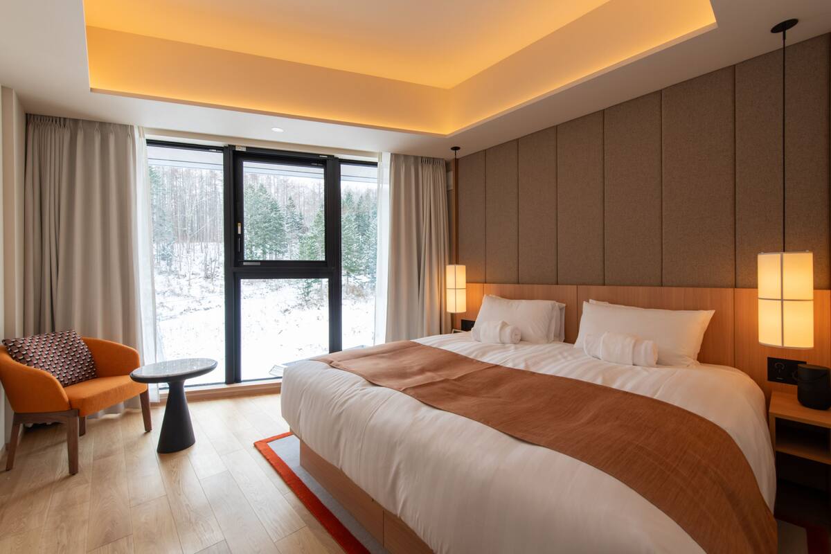 A modern hotel room with a large bed, a small table, a chair, and a view of trees through the window.