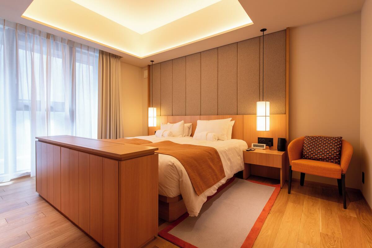 A modern hotel room with a wooden desk, a bed with white linens, a nightstand, and a chair with a patterned cushion.