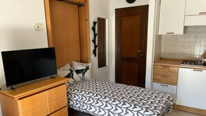 A compact bedroom with a bed, a TV on a wooden dresser, a clock, and a kitchen area with cabinets and a stove. - Cozy Giava's Mountain Place in downtown Aprica (Aprica)