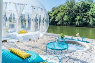 Una cúpula transparente con una cama y una pequeña piscina sobre una terraza de madera junto a un río.