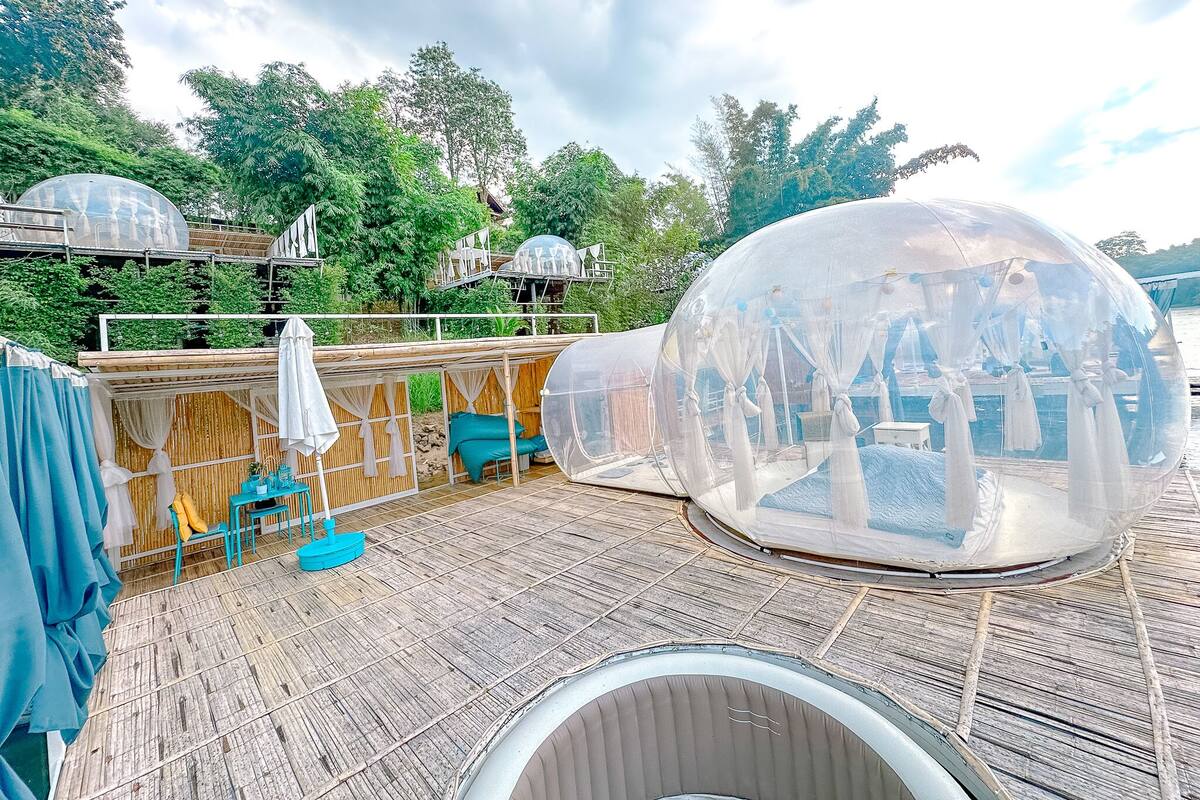A transparent spherical enclosure on a wooden deck with a view of a lush garden and a building in the background.